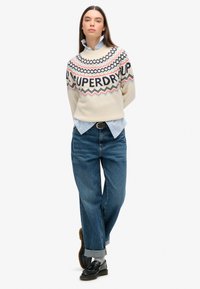 Suéter de punto crema con patrones geométricos multicolores y logo, combinado con camisa azul clara, jeans azules holgados y mocasines negros.