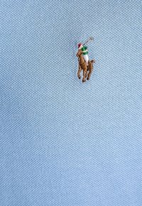 Jasnoniebieska koszulka polo z fakturowanej tkaniny. Na niej haftowany brązowy koń z jeźdźcem ubranym na zielono i biało, w czerwonej czapce.