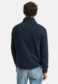 Pullover in fleece blu navy con colletto alto, polsini a costine e una vestibilità rilassata, abbinato a jeans chiari. Texture liscia e design informale.