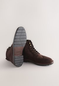 Hnedé semišové členkové čižmy so šnurovaním, zlatými očkami, brogue zdobením a rebrovanou gumovou podrážkou pre lepšiu trakciu.