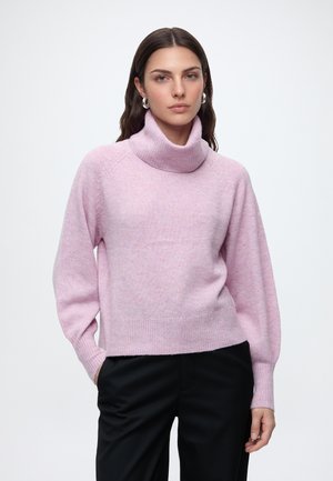 Femme aux cheveux foncés portant des boucles d'oreilles en argent en forme de créoles, un pull col roulé rose clair et un pantalon noir sur un fond blanc uni.