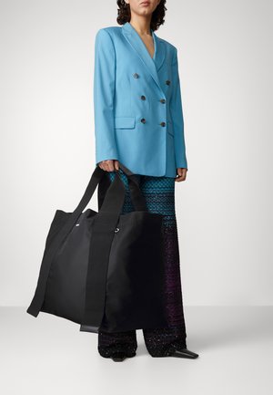 Persoon in een felblauwe blazer en gestreepte brede broek met een grote zwarte schoudertas met lange handvatten.