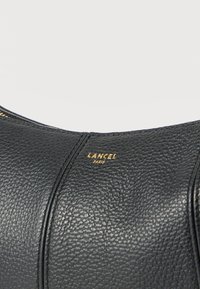 Μαύρη δερμάτινη τσάντα με ανάγλυφη υφή, χρυστό φερμουάρ και το λογότυπο "LANCEL PARIS" ανάγλυφο σε χρυσό στο κέντρο του μπροστινού πίνακα.