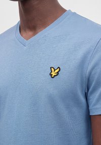 Άνδρας που φοράει ανοιχτό μπλε T-shirt με V-λαιμόκοψη και ένα μικρό κίτρινο σήμα πουλιού στο αριστερό στήθος.