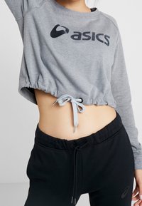 Felpa grigia corta con coulisse in vita e logo "asics" nero, abbinata a pantaloni jogger neri con vita elastica.