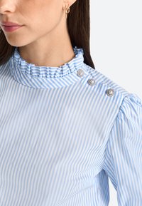 Blusa a rayas azul claro y blanca con un cuello con volantes y tres botones decorativos en el hombro. Tejido suave y mangas abullonadas.
