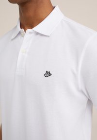 Polo blanco con tejido texturizado, con una tapeta de tres botones y un pequeño logotipo de pájaro bordado en negro en el lado izquierdo del pecho.