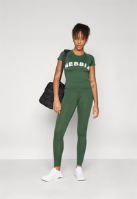 Conjunto de ropa activa verde que consiste en una camiseta ajustada con el logo "NEBBIA" y leggings de cintura alta, combinados con zapatillas blancas y una bolsa negra.