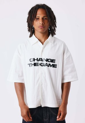 Giovane uomo con dreadlocks che indossa una camicia bianca con "CHANGE THE GAME" stampato in lettere nere in grassetto, in piedi davanti a uno sfondo bianco semplice.