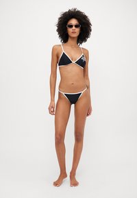 Juodas bikini rinkinys su baltu apvadu, turintis trikampį viršų ir žemai sėdinčius apatinius, pagamintas iš sklandžios medžiagos. Rodytas neutralioje fone.