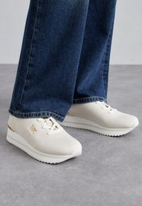 White mesh sneakers with a textured finish, featuring a gold accent and wedge rubber sole, paired with dark blue denim jeans.