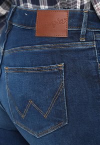 Kék farmernadrág barna bőr Wrangler címkével, sárga varrással és egy hátsó zsebbel, amelynek jellegzetes cikkcakk mintázata van.