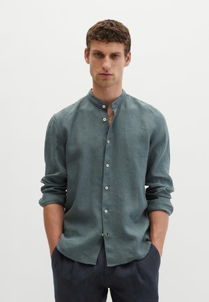 Mężczyzna z krótkimi kręconymi włosami, ubrany w turkusową koszulę z długim rękawem z podwiniętymi rękawami i ciemne spodnie, z rękami w kieszeniach.