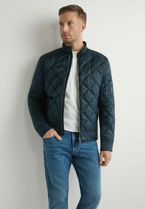 Mężczyzna w granatowej pikowanej kurtce na białej koszuli i niebieskich dżinsach, stojący na tle jasnozabarwionej ściany, patrzący w lewo z ręką w kieszeni.