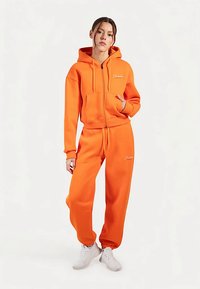 Orange hættetrøje med frontlommer og snoretræk, kombineret med orange træningsbukser med elastiske manchetter. Begge dele har et logo-detalje.