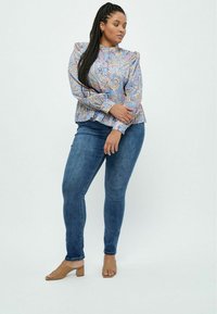 Langærmet, mønstrede bluse i lyseblå med paisleymønster og flæser ved skuldrene, parret med mørkeblå skinny jeans og tan blokhælede sandaler.