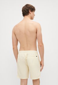 Pantalones de baño amarillo claro con cinturilla elástica, bolsillo trasero y aberturas laterales, mostrados por detrás en una persona con pelo corto.