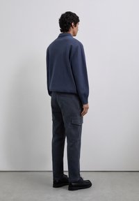 Hombre de pie frente a una pared gris, vistiendo una camiseta de polo de manga larga azul marino, pantalones cargo oscuros y zapatos negros sobre un suelo gris.