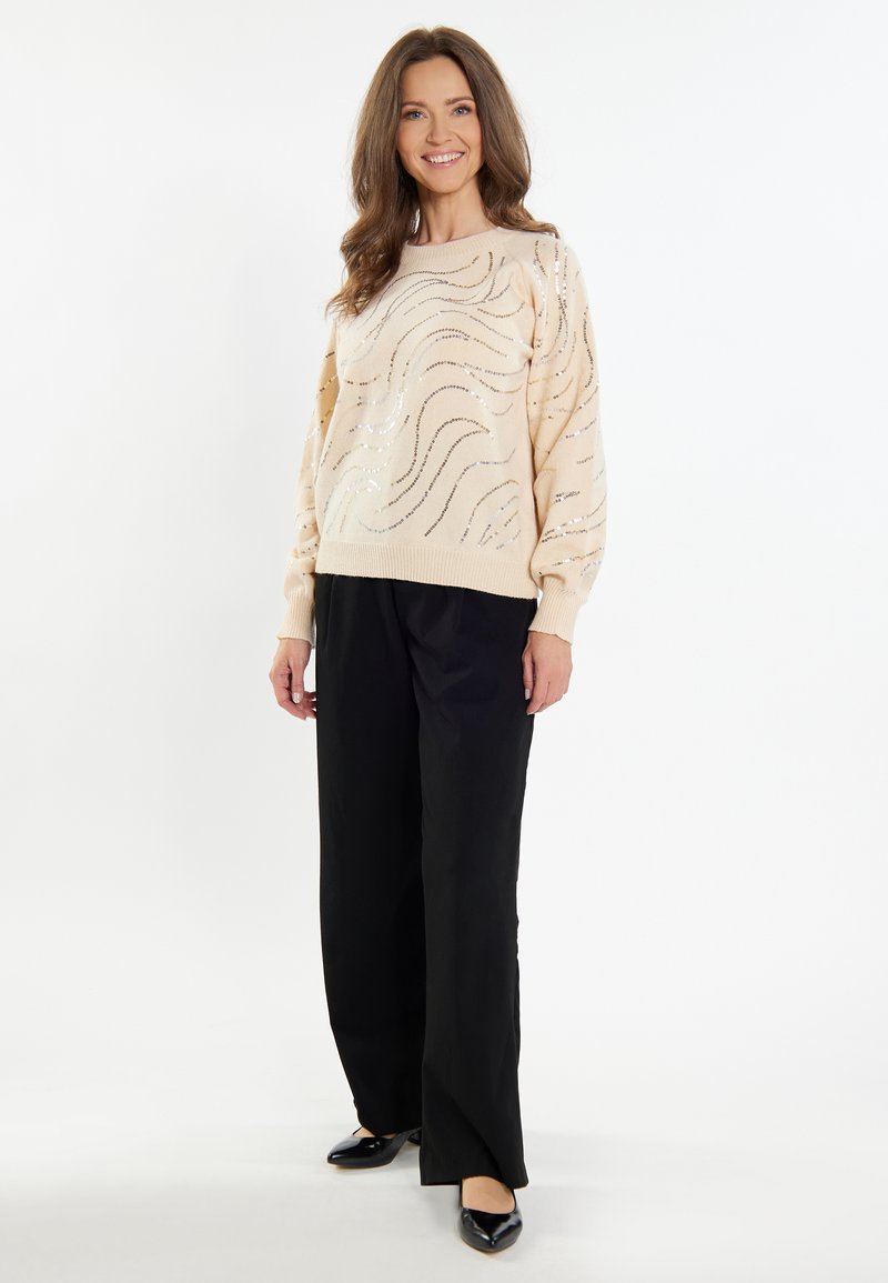 Beige kötött pulóver fémes hullámmintákkal, fekete, széles szárú nadrággal és fekete, hegyes orrú balerinacipővel párosítva. Textúrált kivitelezés, laza fazon.