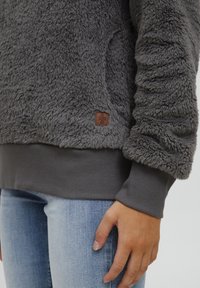 Pullover in felpa grigia con superficie tessurizzata, orlo e polsini a costine. Include una piccola etichetta logo marrone sul lato inferiore.