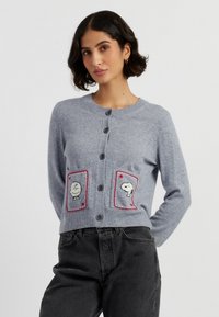 Een vrouw met kort, donker haar modelleert een lichtgrijze cardigan met cartoonpersonages op de zakken, gecombineerd met hoge, zwarte jeans.