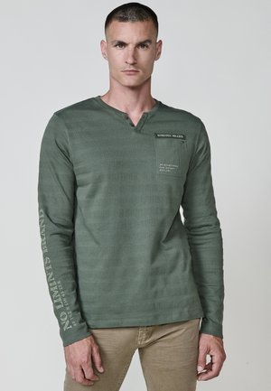 Žalia ilgomis rankovėmis marškiniai su henley iškirpte, tekstūruota juostele, kišenė su logotipo detalėmis ir tekstu kairėje rankovėje.