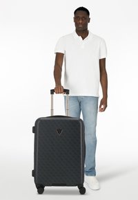 Valise rigide noire avec un motif géométrique, poignée extensible et quatre roues. Elle présente un accent de logo triangulaire sur le devant.