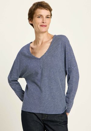 Frau mit kurzen Haaren, die ein blau-weiß gestreiftes Langarmoberteil und dunkle Hosen trägt, steht mit den Händen hinter dem Rücken.