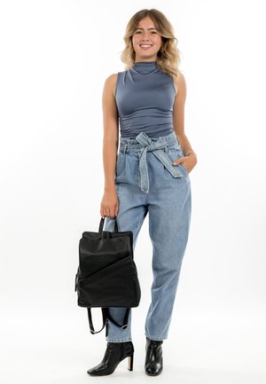 Jonge vrouw die lacht, met een grijs mouwloos topje, hooggetailleerde jeans met ceintuur, zwarte laarzen met hakken, en een zwarte leren rugzak vastgehouden aan het handvat.