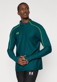 Τιρκουάζ αθλητικό μακρυμάνικο μπλουζάκι με φερμουάρ στο γιακά, που διαθέτει ανακλαστικές κίτρινες λεπτομέρειες και ανάγλυφα σχέδια στα μανίκια.