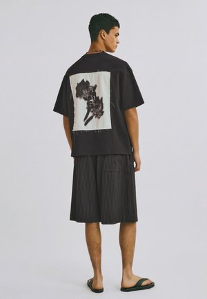 Mand iført sort oversized T-shirt med en stor hvid lap, der viser et mørkt blomsterprint på ryggen, brede shorts med nålestriber og sorte sandaler.