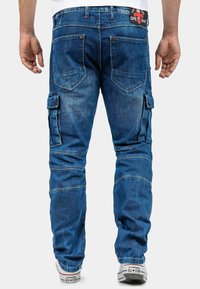 Pantalons cargo en denim bleu moyen avec plusieurs poches, sections délavées et coupe droite. Présente une ceinture de marque et des détails couture.