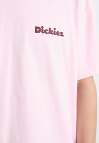 Pink cotton t-shirt featuring a brown "Dickies" logo on the left chest area. Short sleeves and a relaxed fit. Soft texture.