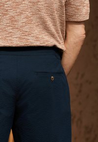 Un pantalon texturé bleu marine présente une poche arrière avec un détail en bouton en laiton. Le tissu a un motif subtil, complété par un haut marron clair.