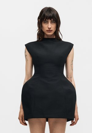 Γυναίκα με σκούρα μαλλιά και τατουάζ, που φοράει ένα δομημένο μαύρο φόρεμα με υπερβολικούς γοφούς και επενδεδυμένους ώμους, στέκεται μπροστά σε ένα απλό λευκό φόντο.