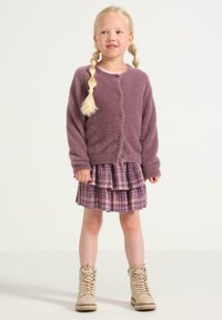 Violetti pörröinen neuletakki, ruudullinen röyhelöhame vaaleanpunaisen ja violetin sävyissä, beige nauhakenkä koristeellisilla yksityiskohdilla.