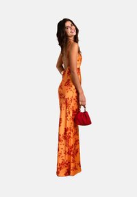 Oranges flieder Maxi-Kleid mit dünnen Trägern und einem tiefen Rücken. Modell hält eine rote Umhängetasche. Es hat eine glatte Textur und eine schlanke Silhouette.