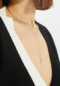 Srebrny naszyjnik z łańcuszka o delikatnym wzorze, z zawieszką, noszony na czarno-białym, teksturowanym ubraniu.