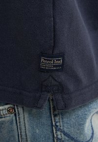 Námořnická modrá bavlněná košile s texturovanou tkaninou a malou cedulkou "Petrol Ind" na lemu, spárovaná se světle modrými džínami.