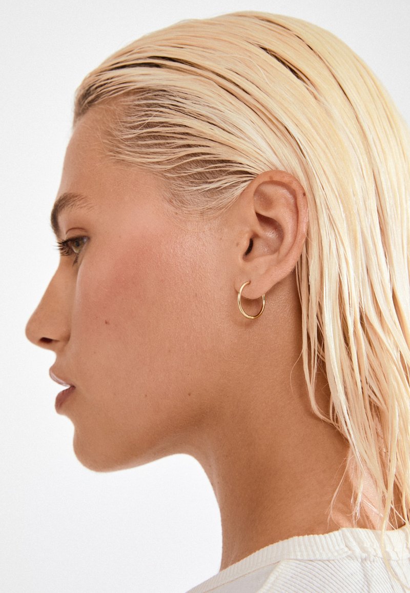 Boucle d'oreille en or affichée sur une oreille. Le design est circulaire et lisse, réfléchissant la lumière subtilement. Les cheveux sont tirés en arrière, révélant la boucle.