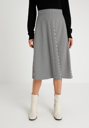 Femme portant un haut noir à manches longues, une jupe midi rayée diagonale en noir et blanc, et des bottines blanches, debout sur un fond uni.