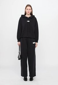 Svart hoodie med en framficka, snören och ett logopatch, kombinerad med vida svarta byxor med en sidopatch. Svarta stövlar och handväska.