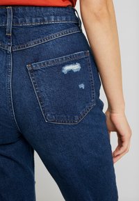 Högmidjade blå denimjeans med en urtvättad bakficka, med subtila slitningar och orange sömnadsdetaljer.
