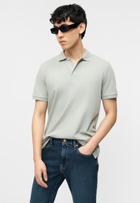 Νεαρός άνδρας που φοράει μαύρα ορθογώνια γυαλιά ηλίου, ανοιχτό γκρι πόλο μπλουζάκι και σκούρα μπλε τζιν, με το ένα χέρι στην τσέπη, κοιτάζοντας προς τα αριστερά.