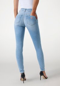 Jean skinny bleu clair avec taille haute, comportant deux poches arrière et un petit logo rouge. Le tissu semble lisse et extensible.