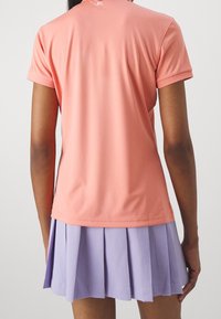 En person som bär en laxrosa kortärmad skjorta och en veckad lavendelkjol, visad bakifrån mot en enfärgad bakgrund.