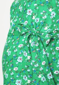 Vestido floral verde com pequenas flores brancas e azuis, detalhe em laço na cintura e um tecido leve com uma textura suave.