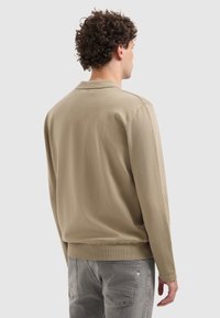 Beige Langarm-Polohemd mit klassischem Kragen, gerippten Bündchen und glatter Textur. Trägt es mit grauen Jeans. Rückansicht.