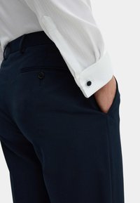 Camicia bianca a maniche lunghe con un motivo testurizzato, abbinata a pantaloni blu scuro. Il polsino della camicia presenta un dettaglio con un bottone nero.