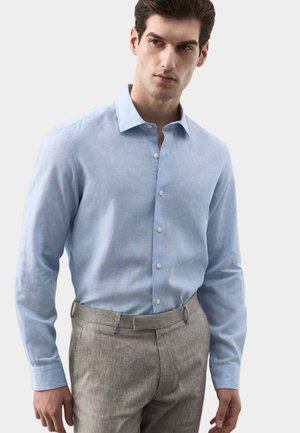 Homme portant une chemise habillée bleu clair boutonnée, rentrée dans un pantalon sur mesure gris clair, debout devant un fond blanc uni.